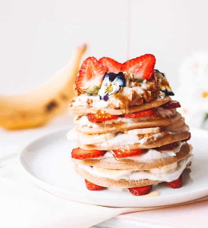 Stack of pancakes layered with strawberry slices and cream, topped with an edible flower and syrup.