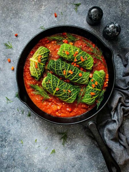 Cast-iron skillet with stuffed cabbage rolls in tomato sauce, garnished with dill and chopped red peppers.
