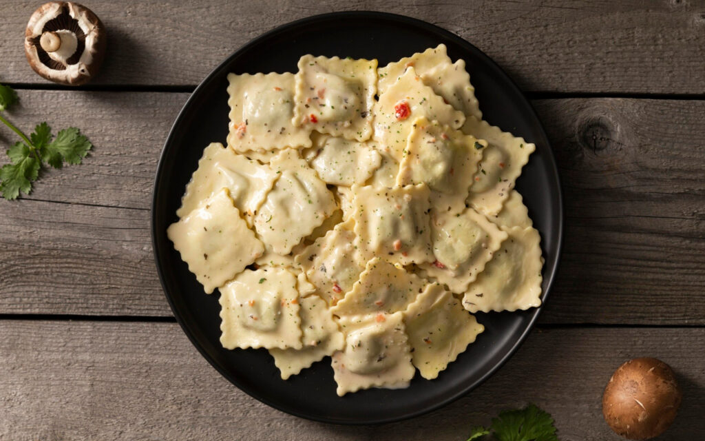 Plate of stuffed cheese ravioli in a light cream sauce on a black plate, set on a rustic wooden table with a mushroom and parsley nearby.