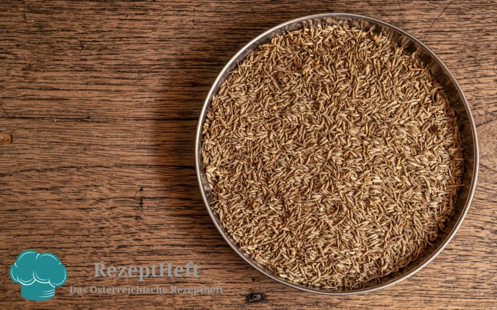 Round metal bowl filled with brown caraway seeds on a wooden surface; RezeptHeft watermark bottom-left.